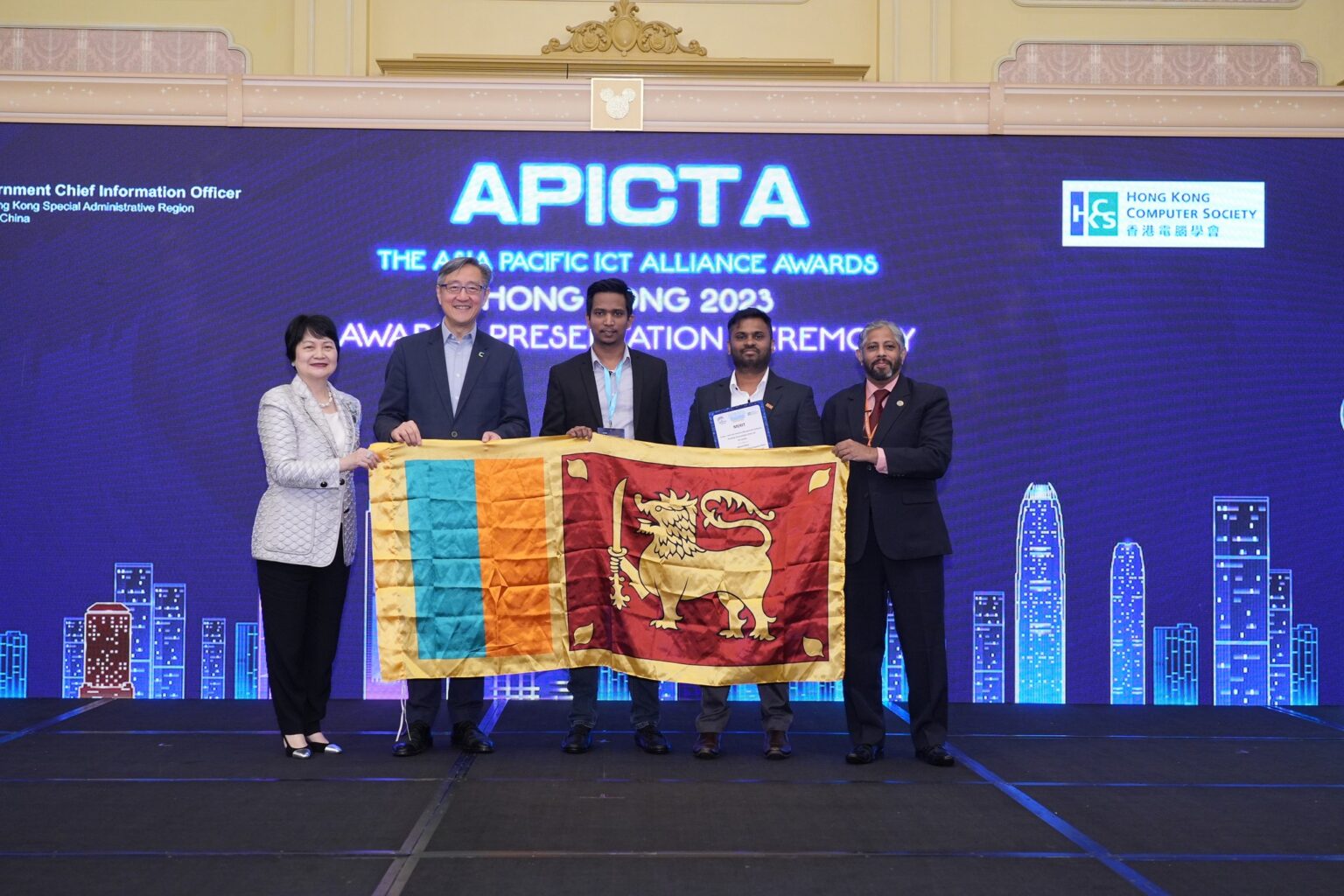 Namal Attanayake and a colleague holding the Sri Lankan flag on stage at the APICTA Awards 2023 in Hong Kong